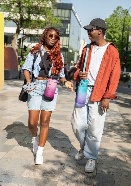 Two people walking through a city, each carrying an EnergyBud half gallon on a shoulder strap